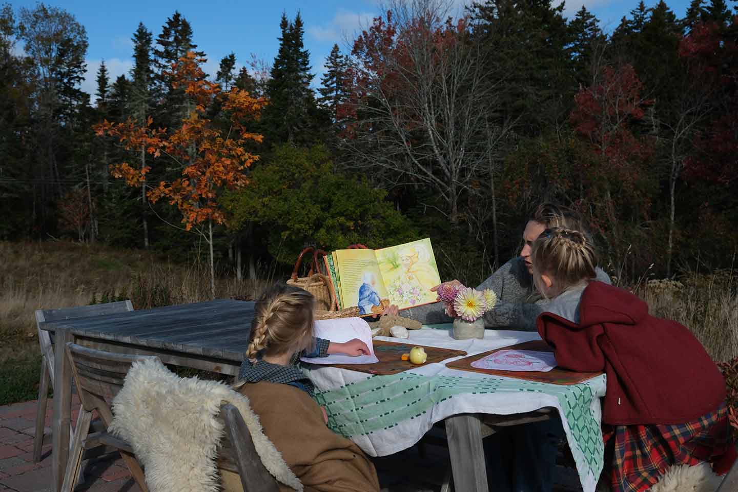 A mother reading a book to her two daughters at a table outside