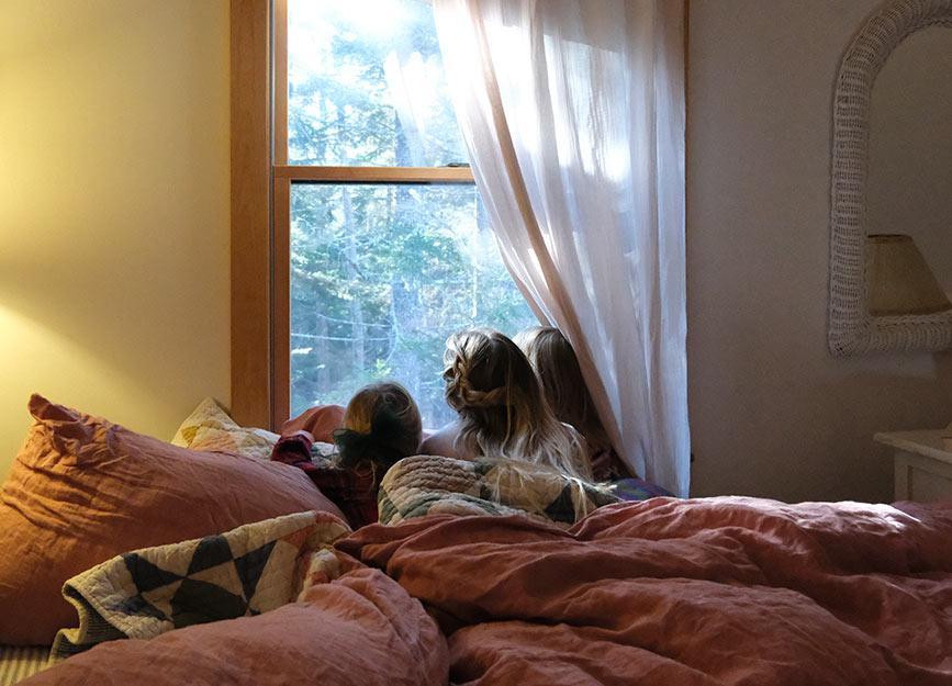 A woman lying in bed looking out the window with her two daughters