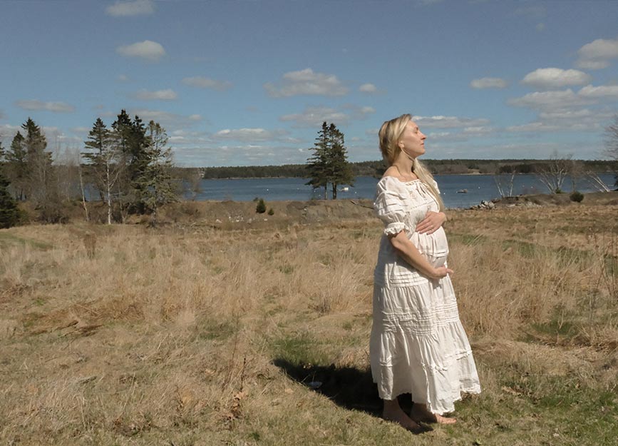A woman in a white cotton dress holding her baby bump in a field